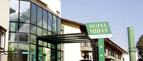 Fachada moderna de hotel con un amplio ventanal, zona de acceso cubierta y un cartel con el nombre y número de contacto.