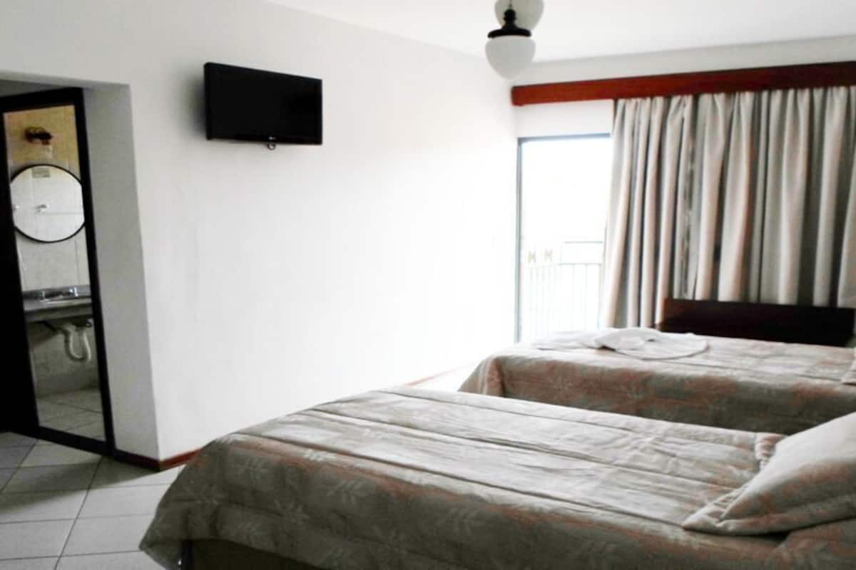 Habitación de hotel con dos camas, televisión, baño con espejo y ventana con cortinas.