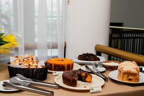 A buffet table with various baked goods including a cake labeled "Bolo Milho" and another labeled "Bolo Cenoura". - Royal Inn Hotel (Cornelio Procopio)