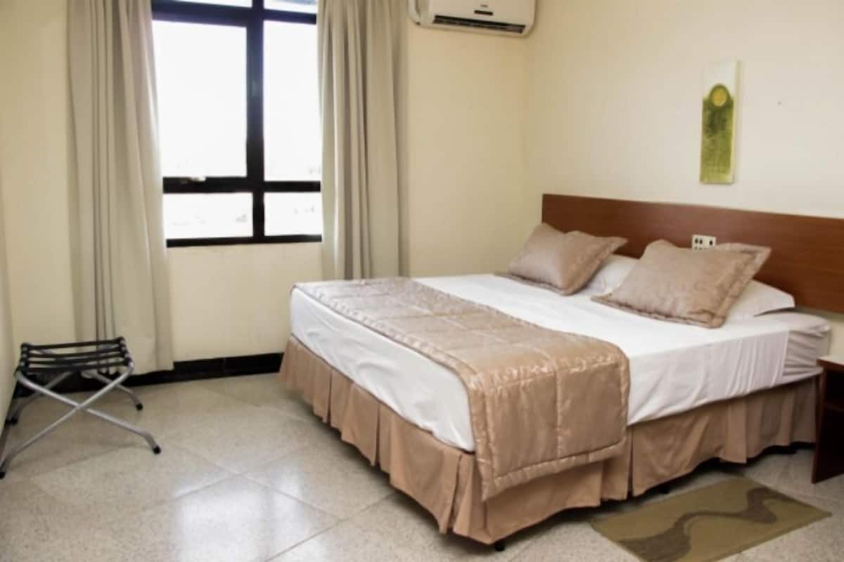 Una habitación de hotel con cama, cortinas, un equipo de aire acondicionado y un escritorio pequeño.