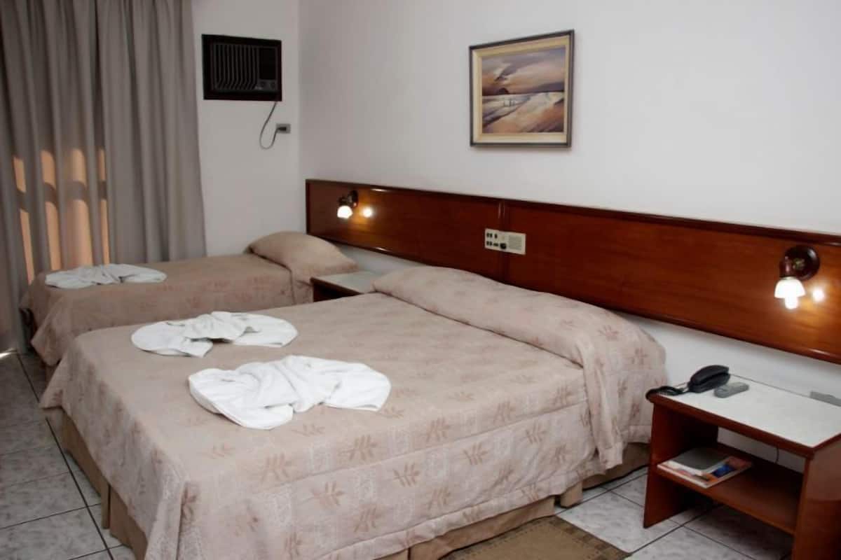 Habitación de hotel con dos camas, una mesita de noche con lámpara, un cuadro en la pared y una ventana con cortinas.