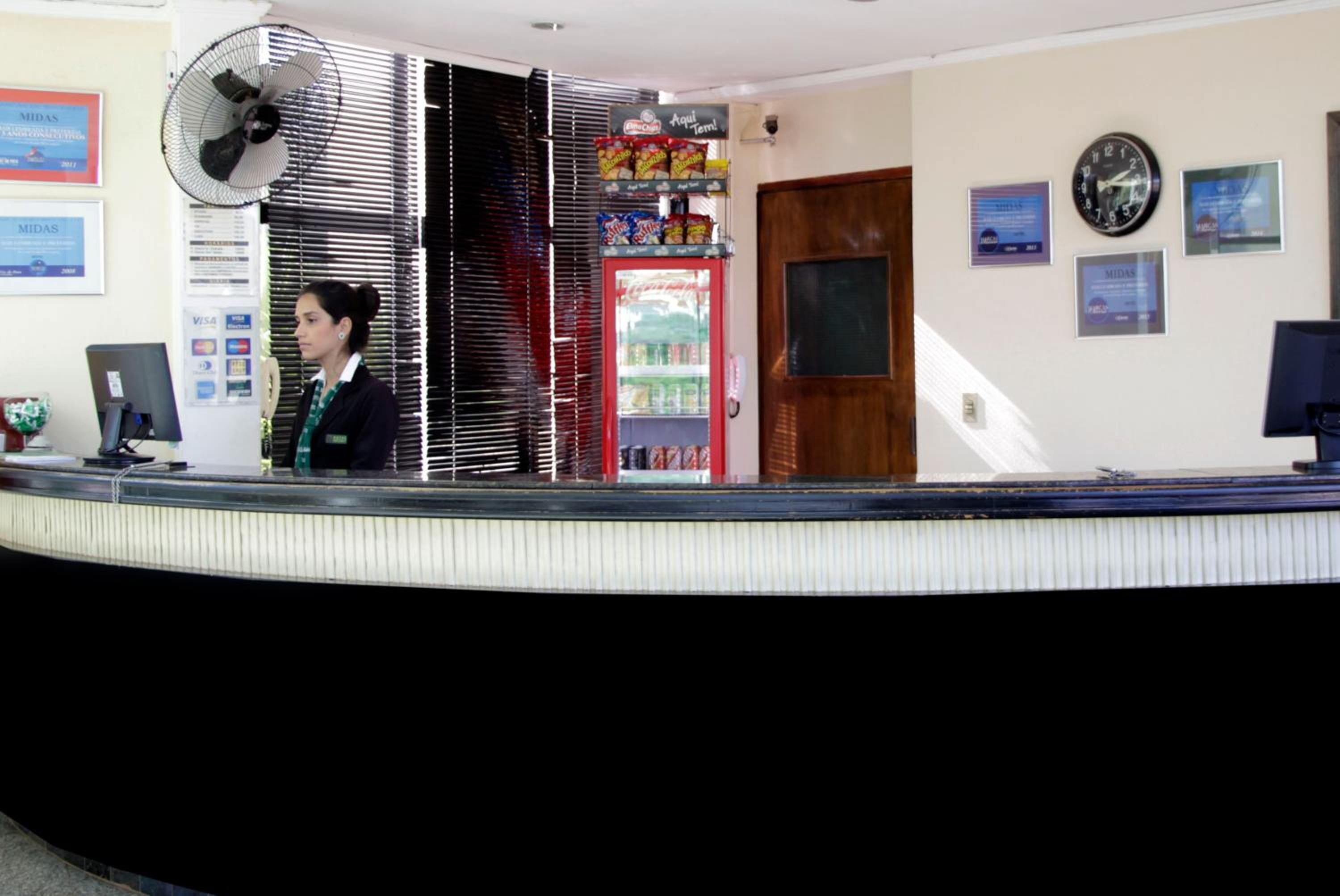 Un'area reception con un bancone, una ventola, un frigorifero fornito di bevande e una sveglia appesa al muro.