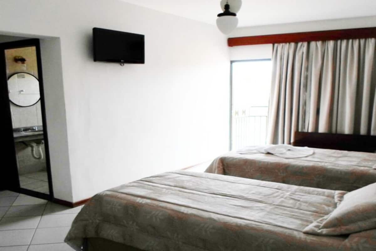 Habitación de hotel con dos camas, un televisor montado en la pared, un baño con lavamanos y espejo, y una ventana con cortinas.