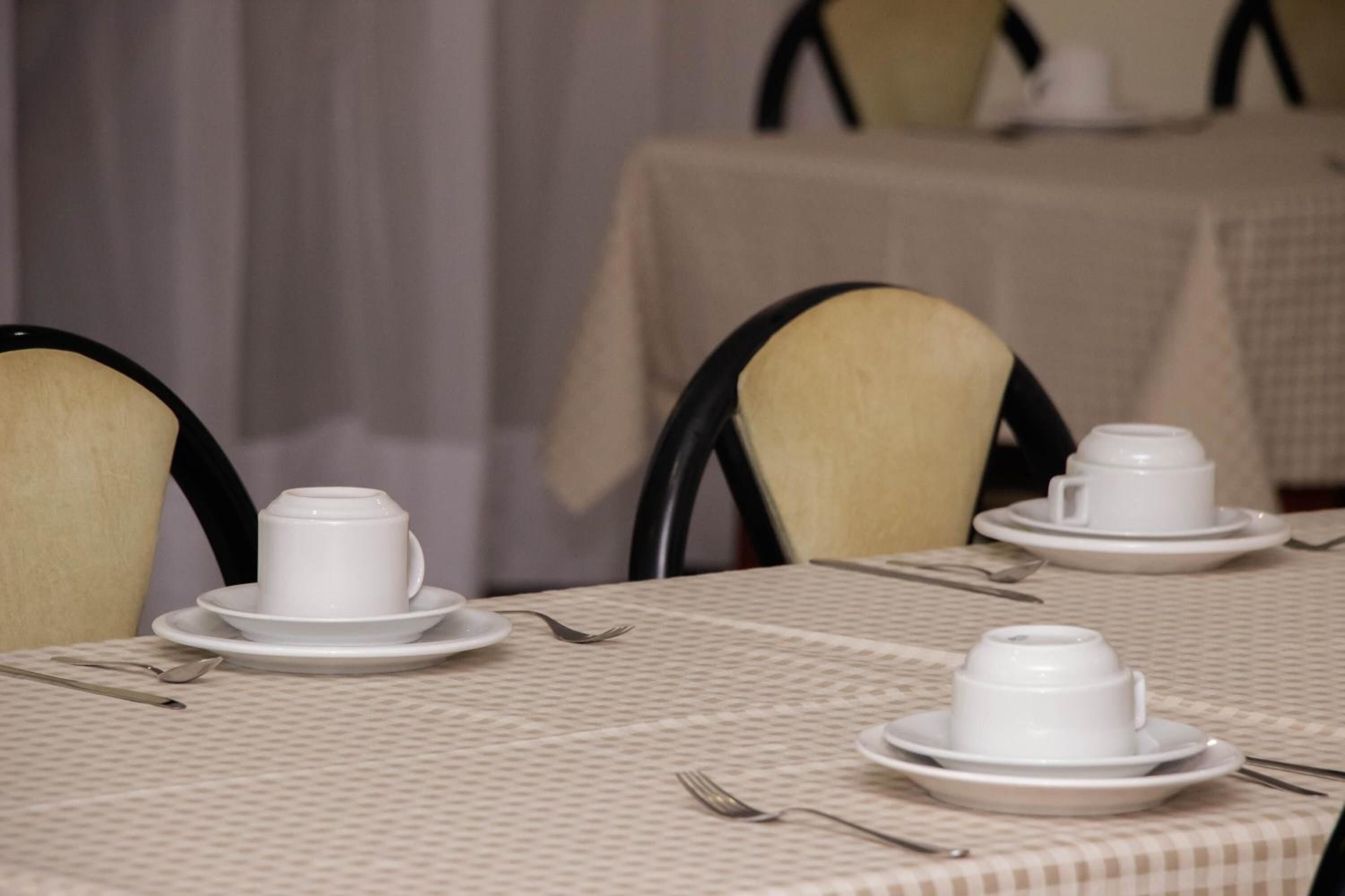 Una mesa dispuesta con tazas y platillos blancos, cubiertos y un mantel a cuadros.