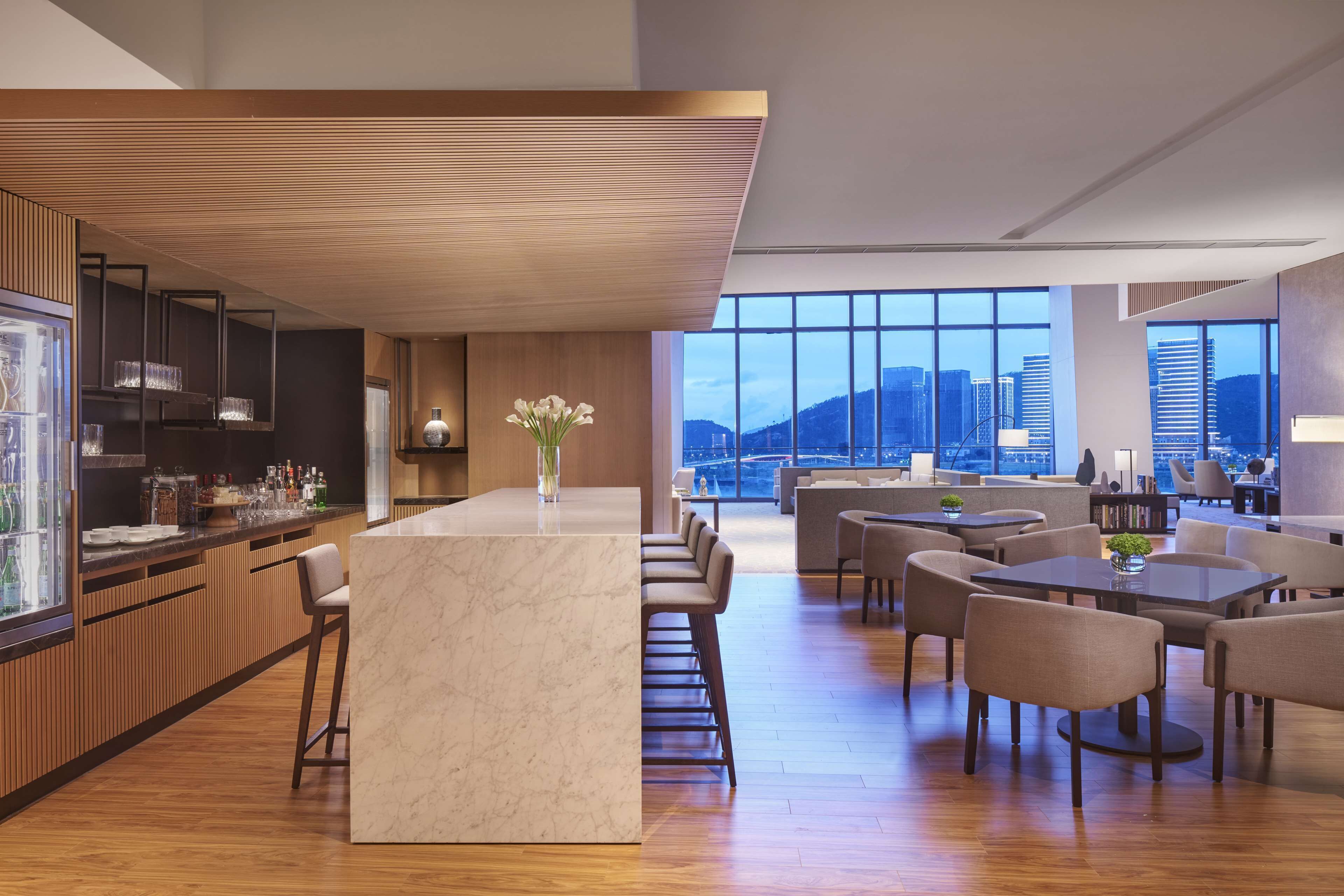 Un hall d’hôtel moderne doté d’un bar, de différents espaces de détente et de grandes fenêtres offrant une vue sur la ville.