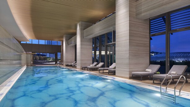 An indoor swimming pool with clear blue water, surrounded by lounge chairs and large windows offering views of the cityscape at night. - Hyatt Regency Hengqin (Zhuhai)