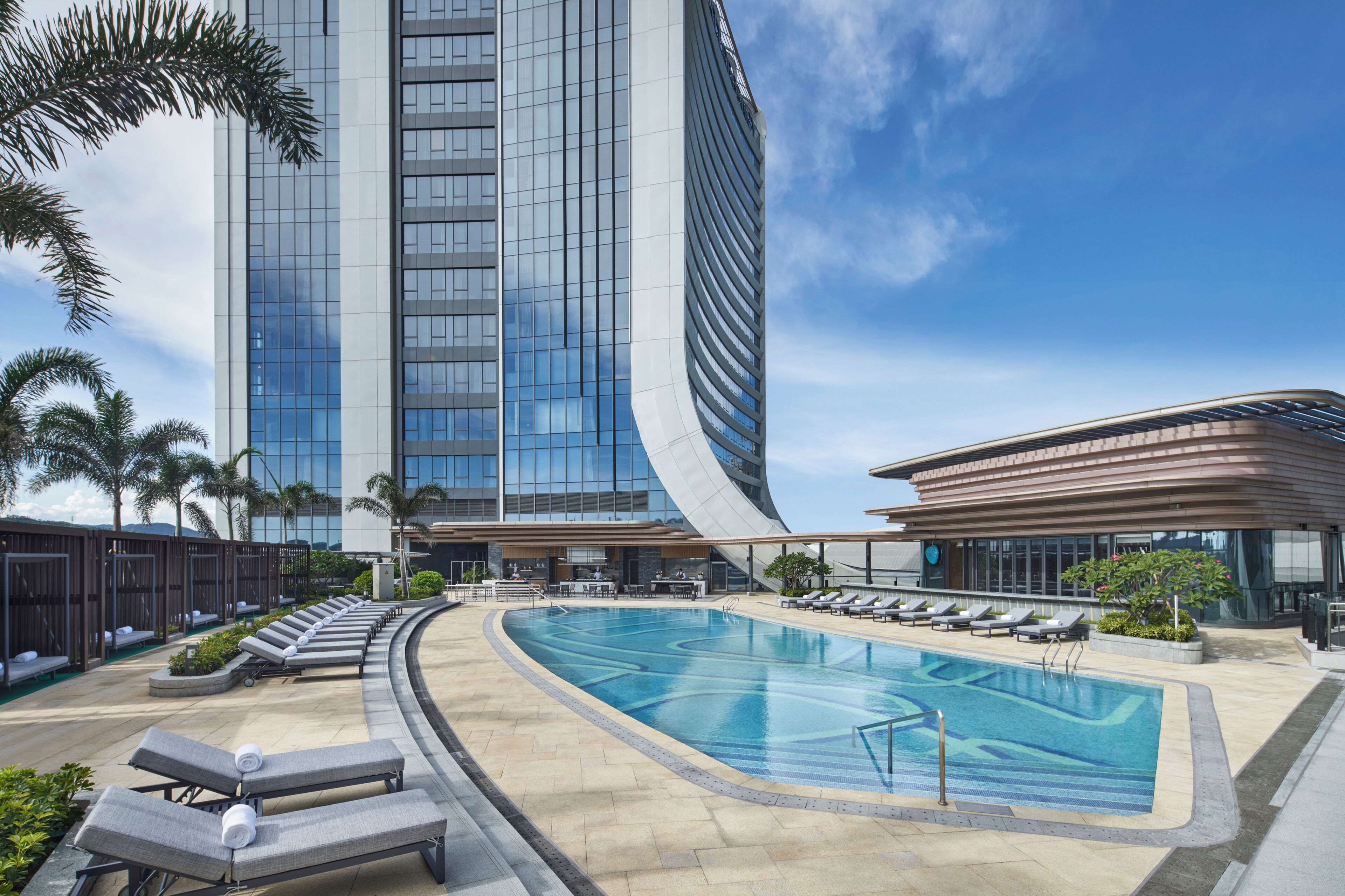 A modern hotel with a curved glass facade, a swimming pool with lounge chairs, and palm trees.