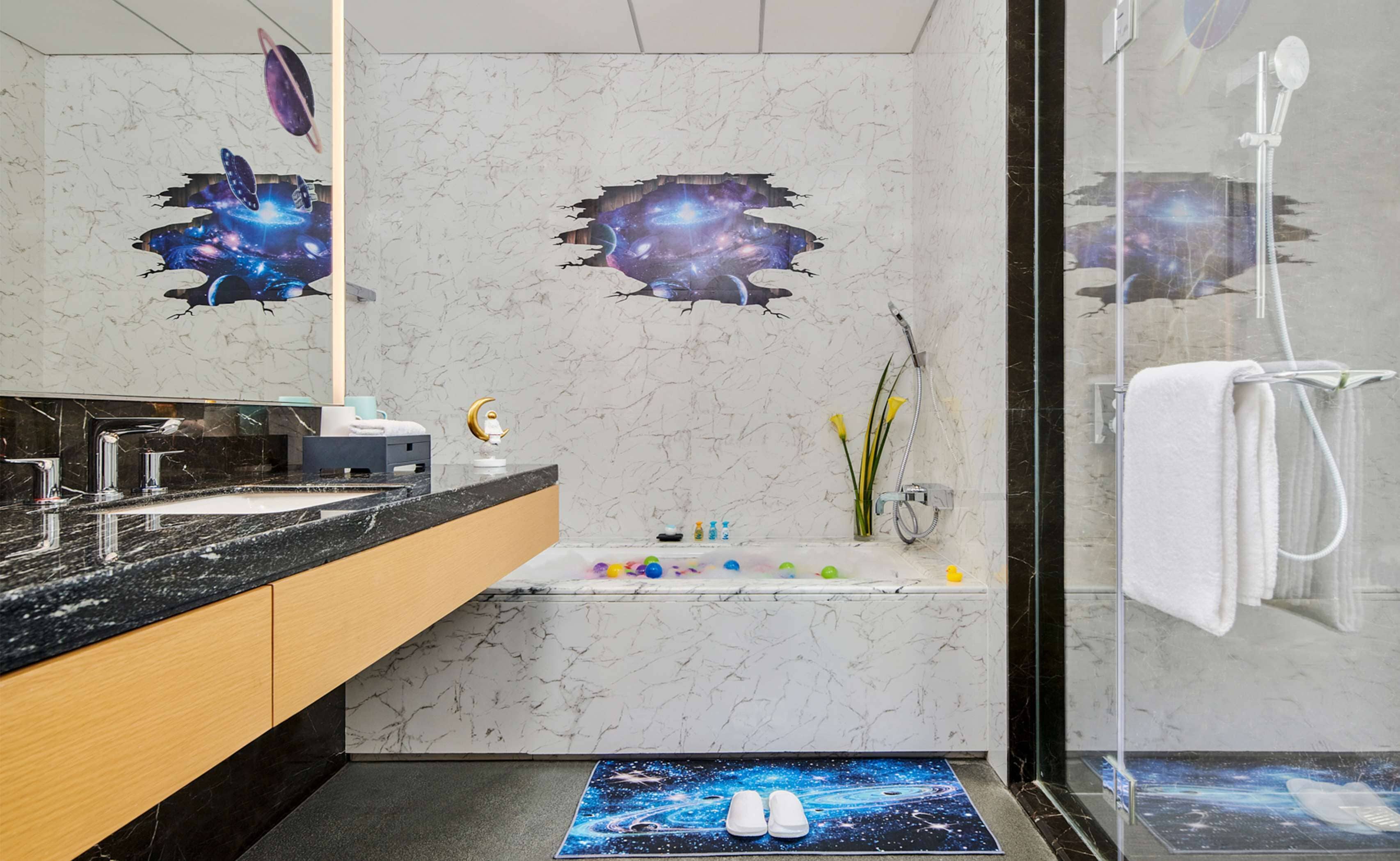Une salle de bain moderne avec un motif mural représentant une galaxie, une baignoire, une douche et un meuble-lavabo doté d’un plan de travail en marbre.