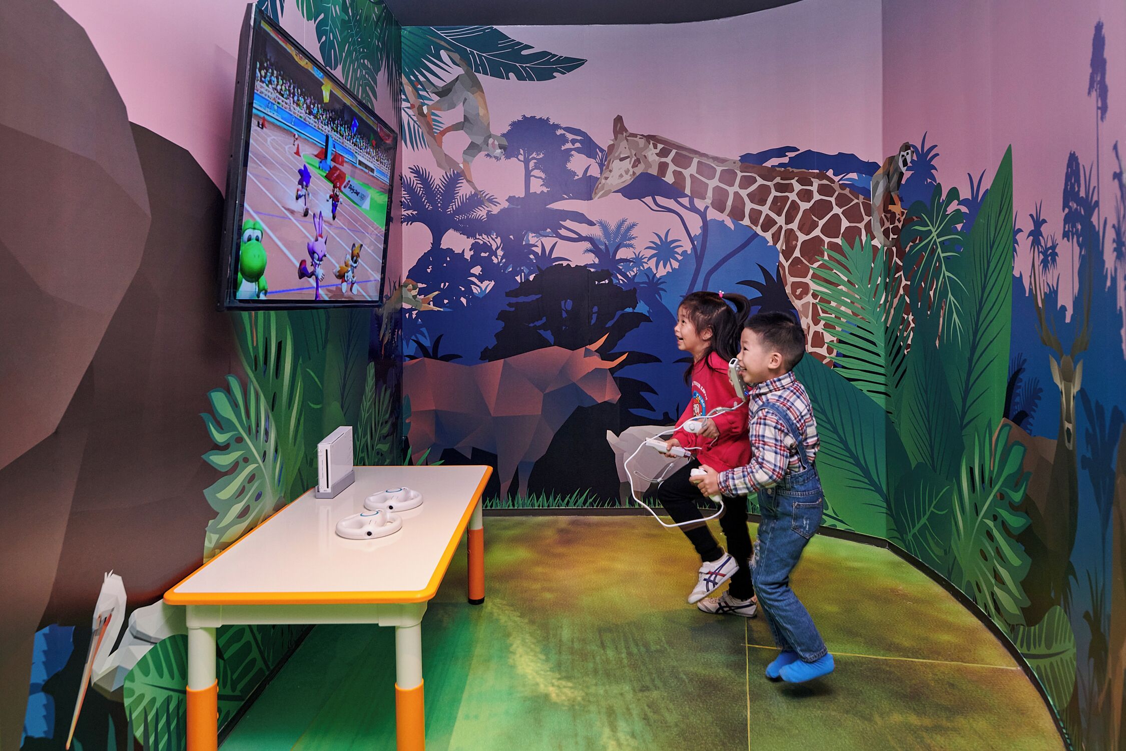 Two children playing a video game in a room with a jungle theme, including a large giraffe and monkey mural.