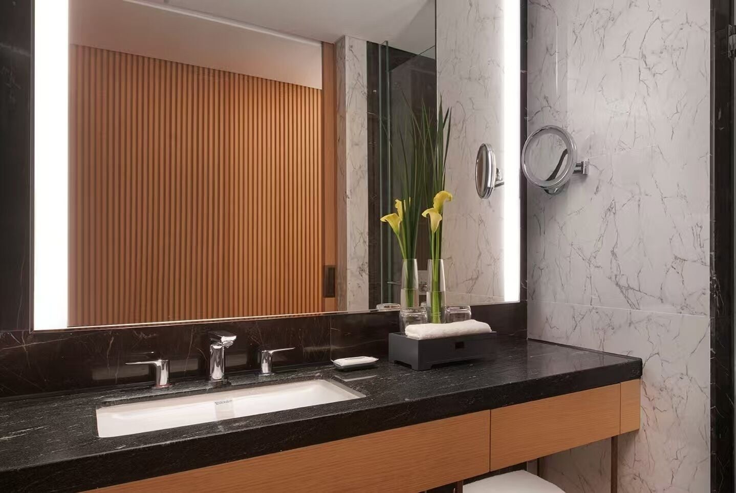 A modern bathroom with a black marble countertop, a large mirror, and a wooden panel wall.