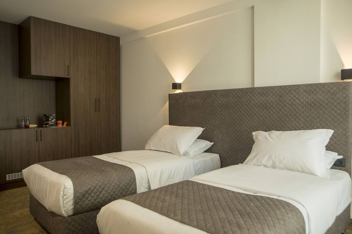 Quarto de hotel com duas camas de solteiro, um guarda-roupa e uma luminária fixa na parede.