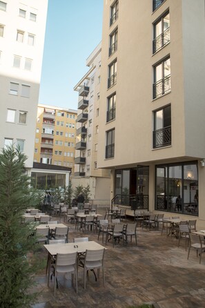 Un área de café al aire libre con mesas y sillas, rodeada de edificios con balcones y ventanas amplias.