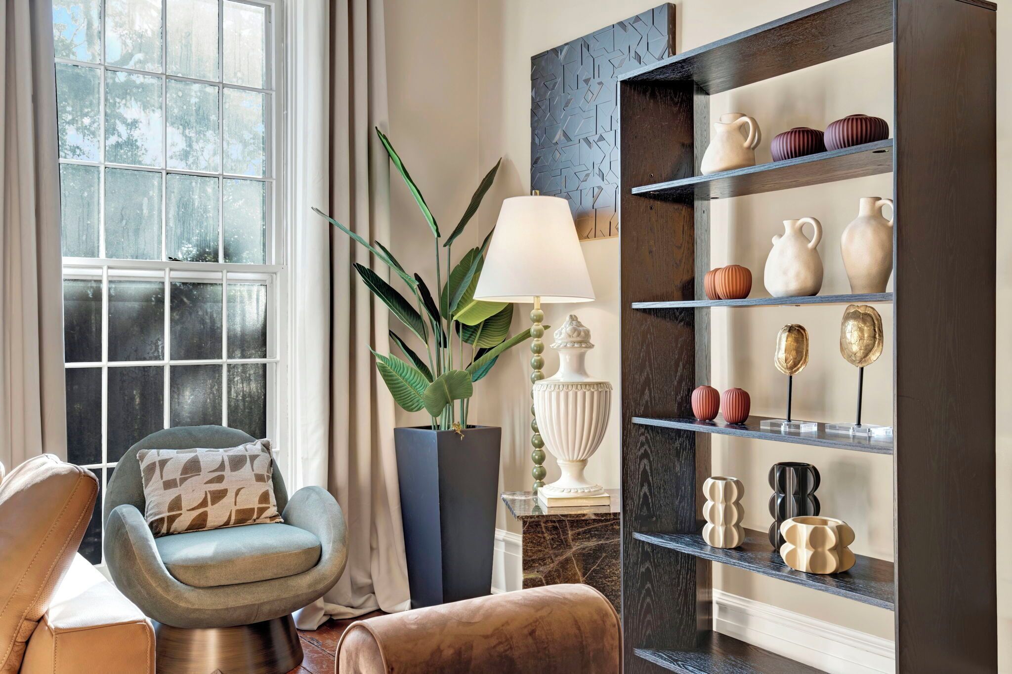 Una sala de estar acogedora con un sillón cómodo, una planta en maceta, una lámpara y una estantería decorativa con cerámica.