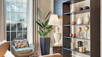 Una sala de estar acogedora con un sillón cómodo, una planta en maceta, una lámpara y una estantería decorativa con cerámica.