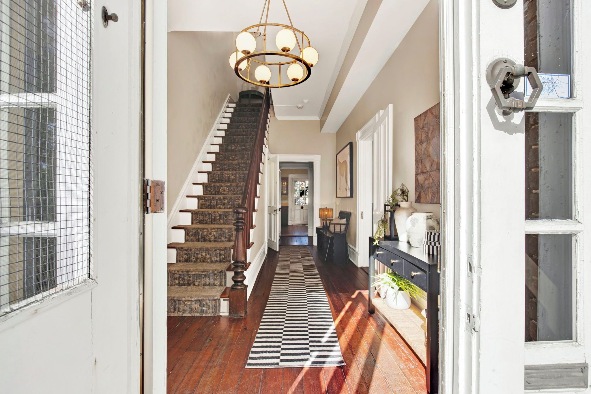 Un pasillo con escalera de madera, candelabro y alfombra estampada.