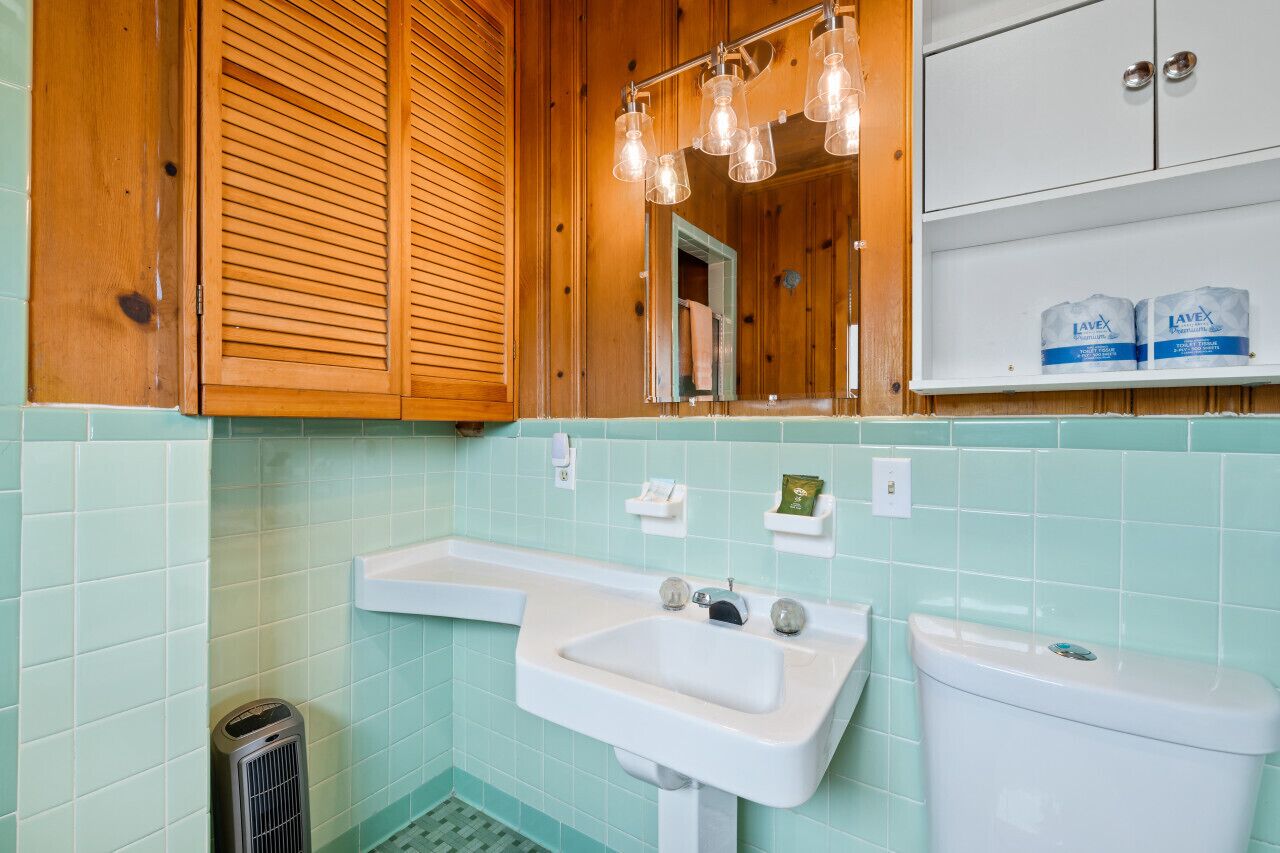 Een badkamer met een wit wastafel, toilet en houten kastjes. Boven het wastafel hangen twee lampen en boven de kastjes hangt een spiegel.