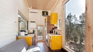 Une petite cabane comprenant un coin cuisine, un canapé et une grande fenêtre donnant sur la forêt environnante.