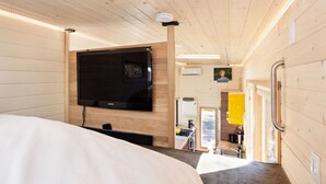 Un intérieur moderne avec un grand téléviseur à écran plat fixé au mur, un lit doté d’une tête de lit et un coin cuisine visible à travers une porte ouverte.