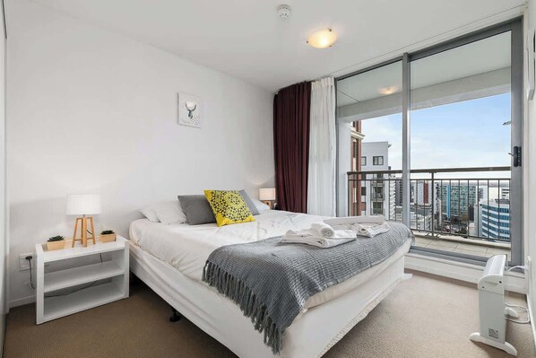 Ein modernes Hotelzimmer mit einem großen Bett, einem Nachttisch mit Lampe, einer kleinen Pflanze und Blick auf die Stadt durch ein großes Fenster.