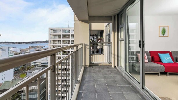 A balcony with a view of the sea, a red sofa, and a glass door leading to a room. - Wonderful Apt in CBD Neighbourhood I Zodiak Stays (Auckland)