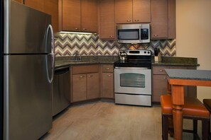 Compact kitchen with modern appliances, a stainless steel refrigerator, oven, and sink, along with a wooden table and chair.
