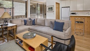 Una sala moderna con un sofá color beige, una mesa de centro de madera y una silla negra. Hay una zona de cocina con armarios blancos y un microondas.