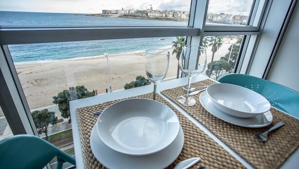 A dining table with white plates and a glass of water, set on a woven placemat, overlooking a beach and the sea.