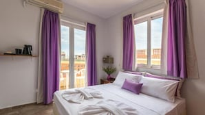 A hotel room with a bed, purple curtains, a window, and a view of buildings. - Maria Hotel (Agios Nikolaos)