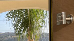 A balcony with a view of hills and a palm tree, a small table with a plant, and a bed with towels. - Maria Hotel (Agios Nikolaos)