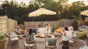 Un grupo de personas sentadas alrededor de un brasero en un patio al aire libre con muebles de mimbre y sombrillas.