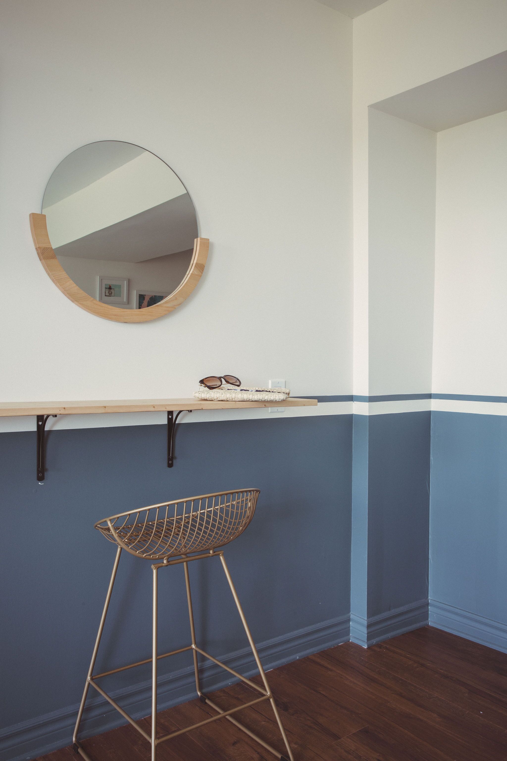 A room with a wooden bar stool, a round mirror, and a shelf with sunglasses and a basket.