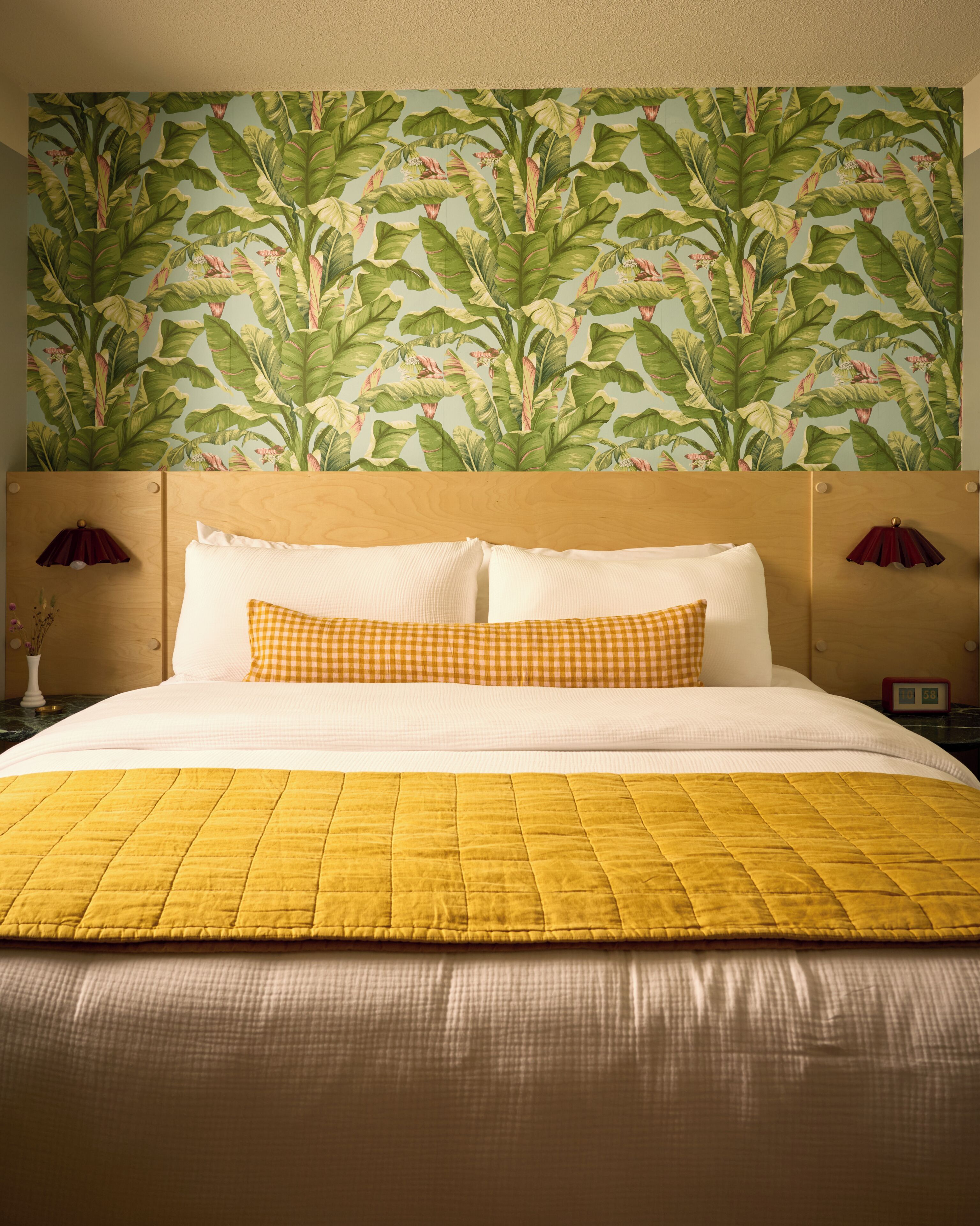 Une chambre d’hôtel avec un grand lit, une tête de lit recouverte d’un papier peint à motifs de feuilles et une table de chevet avec une lampe rouge.