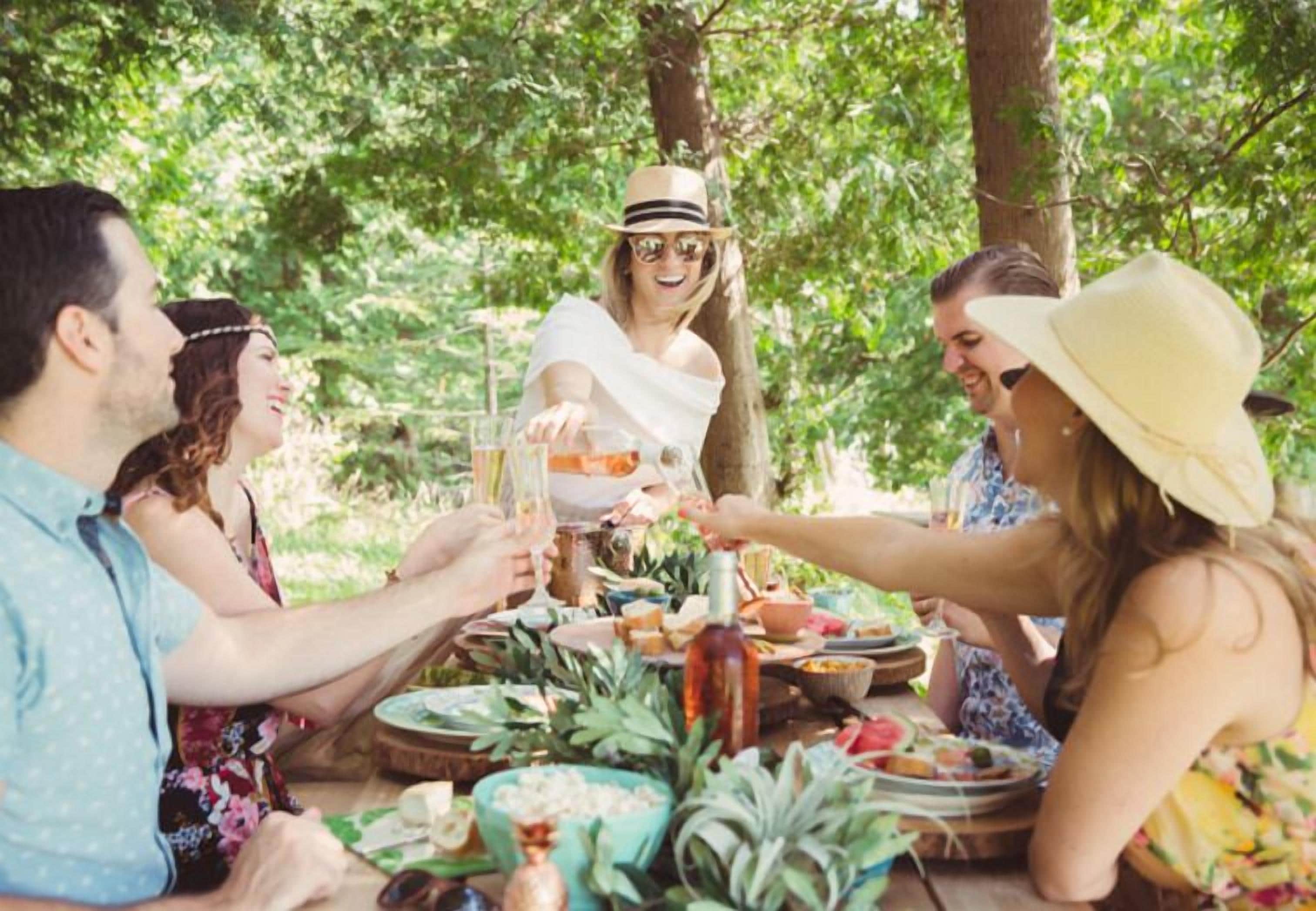 Een groep mensen zit aan een buitentafel met eten en drinken, omringd door groen.
