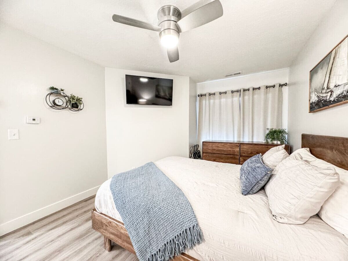 Une chambre à coucher avec un lit, un ventilateur de plafond, un téléviseur fixé au mur et une fenêtre avec des rideaux.