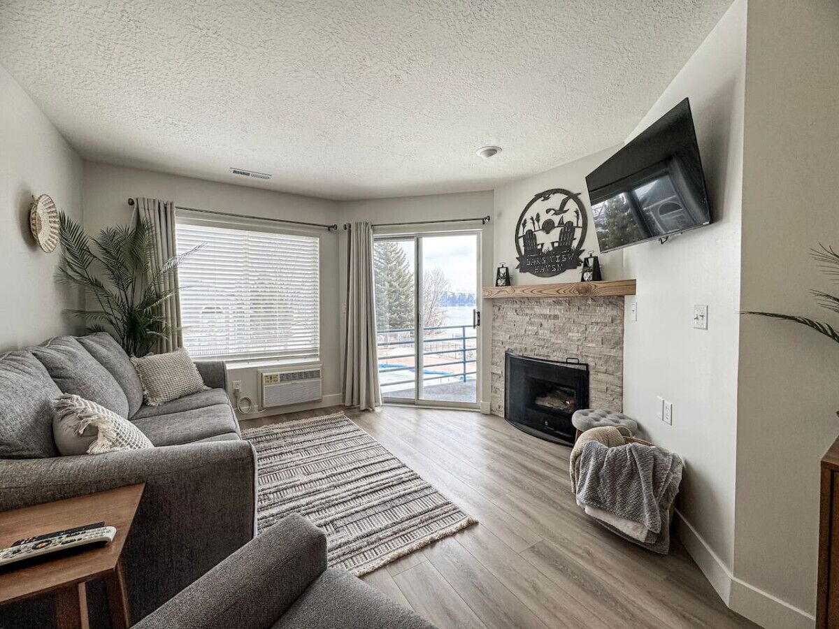 A living room with a grey sofa, a fireplace, a TV mounted on the wall, and a large window with curtains.