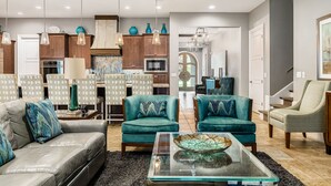 A modern living room with a sofa, two armchairs, a coffee table, and a kitchen in the background. - Built For Vacation | South-Facing, Large Screen Pool, Splash Pad, Game,Movie Room (Four Corners)
