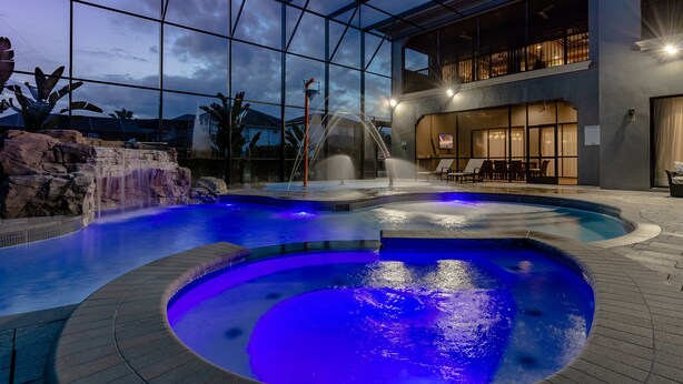 A swimming pool with a waterfall feature, surrounded by a glass enclosure, and a second-story balcony with outdoor seating. - Built For Vacation | South-Facing, Large Screen Pool, Splash Pad, Game,Movie Room (Four Corners)
