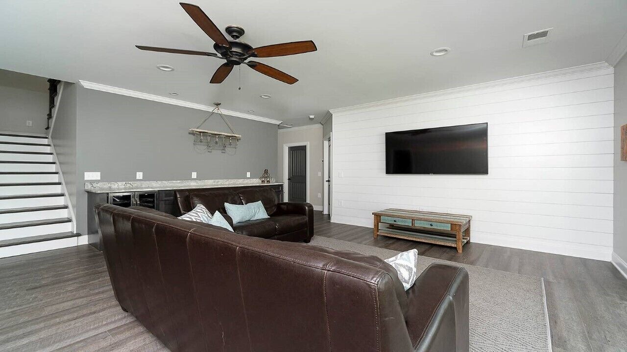 A modern living room with a brown leather sectional sofa, a ceiling fan, a flat-screen TV mounted on the wall, and a kitchen area in the background.