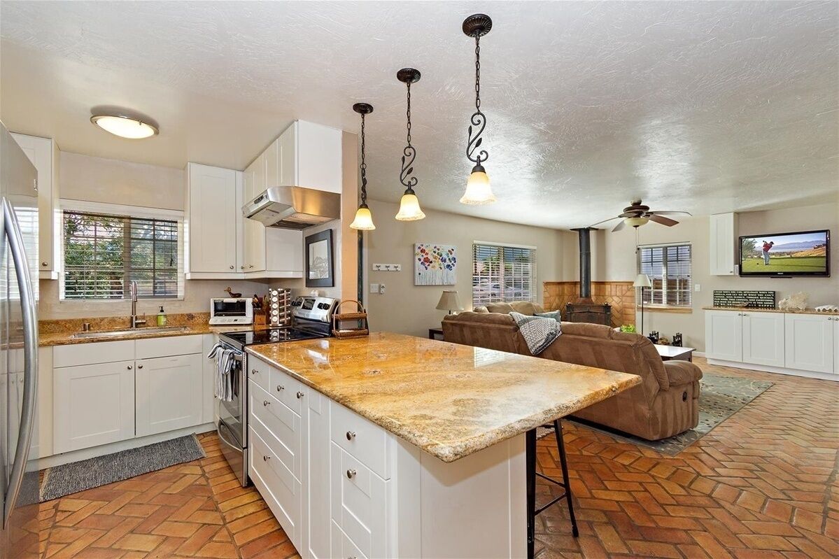 A spacious kitchen with a large island, white cabinetry, and a stone countertop. There is a dining area with a brown leather couch and a television.