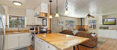 A spacious kitchen with a large island, white cabinetry, and a stone countertop. There is a dining area with a brown leather couch and a television.