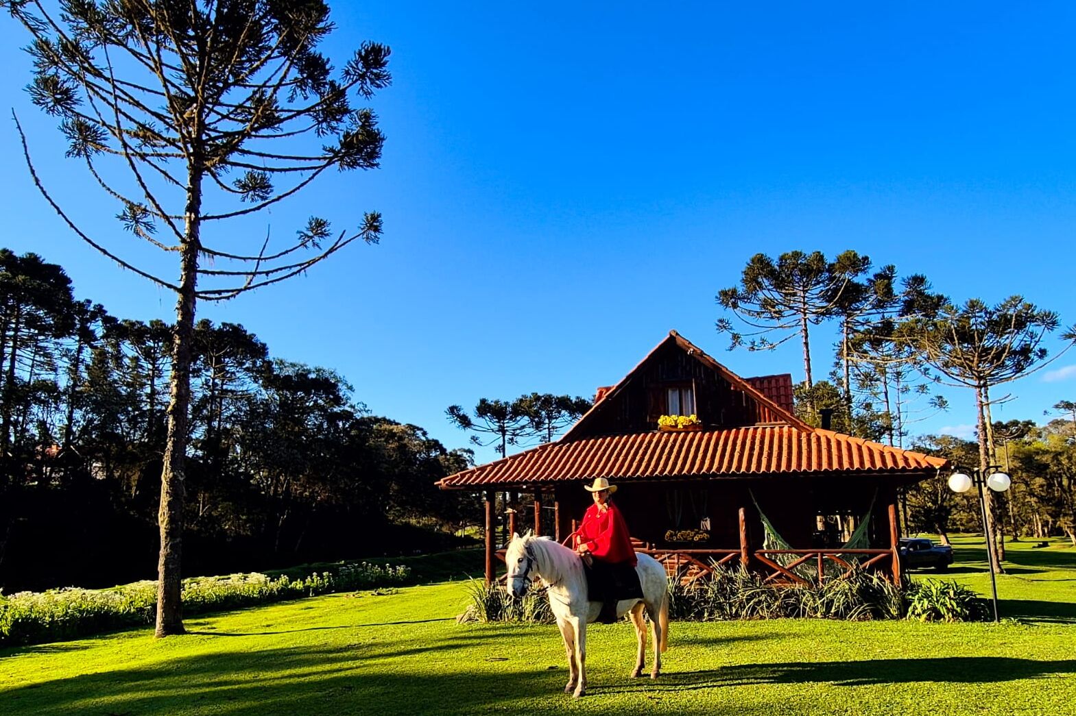 A person on a horse in front of a rustic wooden house with a red roof, surrounded by lush greenery and tall trees.