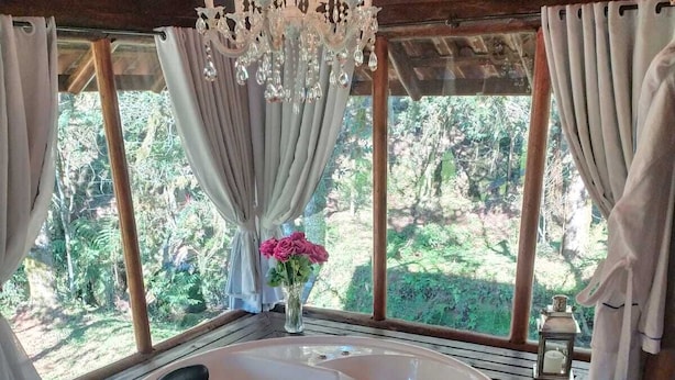 Un bain à remous installé sur une terrasse en bois, agrémenté d’un lustre, de rideaux et offrant une vue sur la verdure.