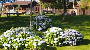 Une maison rustique en bois, avec un toit recouvert de tuiles rouges, entourée d’une végétation luxuriante et de plantes en fleurs.