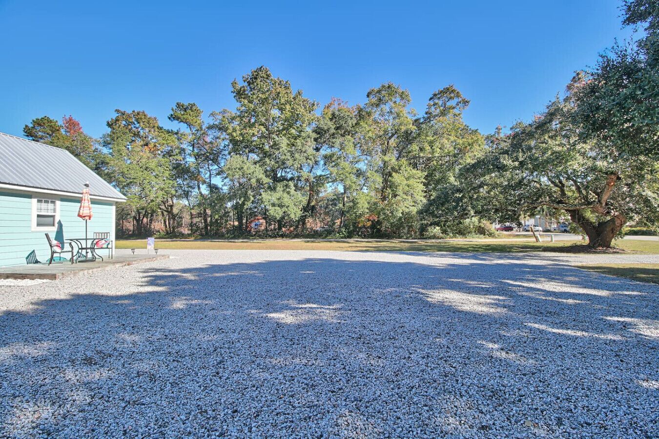 A gravel parking area with a building on the left, outdoor seating, and trees in the background.