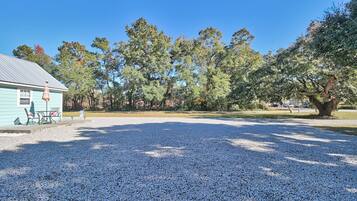 A gravel parking area with a building on the left, outdoor seating, and trees in the background.