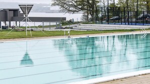 Una piscina con carriles, un edificio moderno al fondo y un área cercada donde personas hacen ejercicio.