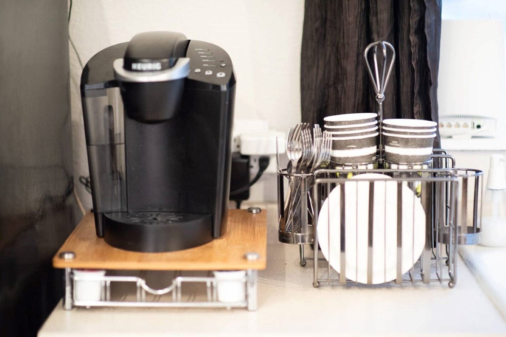 A coffee maker on a wooden tray, a metal utensil holder with plates, and a window with curtains in the background.