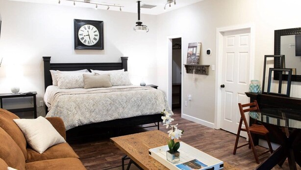 Une chambre à coucher comprenant un lit, une table de chevet, une table basse en bois, une chaise, un ventilateur de plafond, une horloge et une porte.