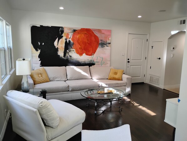 A living room with a sofa, armchair, and coffee table, a large abstract painting on the wall, and a door in the background.