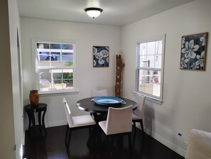 A dining area with a round table, four chairs, two windows, and wall art.
